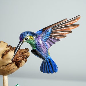 May include: A hand-carved wooden hummingbird figurine featuring vibrant blue, purple, and green painted feathers. The bird is depicted in flight with wings extended, hovering over a textured, light brown wooden flower blossom on a thin stem.