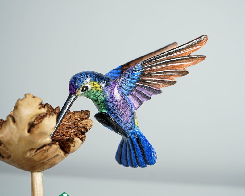 May include: A hand-carved wooden hummingbird figurine featuring vibrant blue, purple, and green painted feathers. The bird is depicted in flight with wings extended, hovering over a textured, light brown wooden flower blossom on a thin stem.