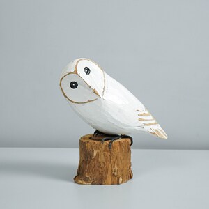 K&ouml;nnte beinhalten: Eine wei&szlig;e Holz-Eulenfigur mit schwarzen Augen und goldenem Schnabel, die auf einem kleinen, braunen Holzstumpf sitzt. Die Eule hat einen leicht geneigten Kopf. Die Figur ist handgeschnitzt und bemalt.
