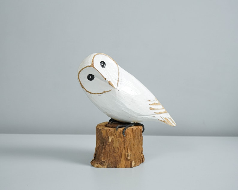 K&ouml;nnte beinhalten: Eine wei&szlig;e Holz-Eulenfigur mit schwarzen Augen und goldenem Schnabel, die auf einem kleinen, braunen Holzstumpf sitzt. Die Eule hat einen leicht geneigten Kopf. Die Figur ist handgeschnitzt und bemalt.