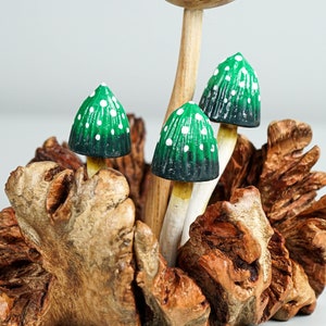May include: A decorative sculpture featuring three small mushrooms with green and black caps adorned with white dots, mounted on a textured, natural brown wooden base. The mushroom stems are light-colored and slender, emerging from the center of the wood.