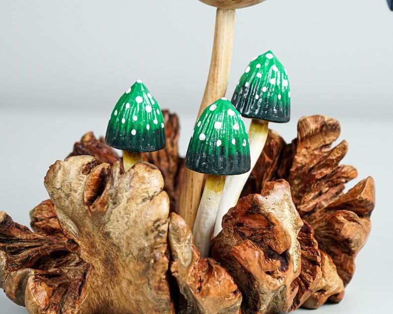 May include: A decorative sculpture featuring three small mushrooms with green and black caps adorned with white dots, mounted on a textured, natural brown wooden base. The mushroom stems are light-colored and slender, emerging from the center of the wood.