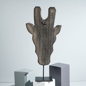 May include: Wooden giraffe head sculpture with a distressed black finish, mounted on a black metal stand. The sculpture is on a white and gray platform.