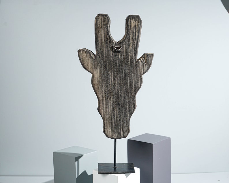 May include: Wooden giraffe head sculpture with a distressed black finish, mounted on a black metal stand. The sculpture is on a white and gray platform.