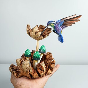 May include: A hand-carved wooden sculpture featuring a vibrant hummingbird with blue, purple, and green plumage hovering over a textured wooden flower, accompanied by three small green mushrooms with white spots emerging from a gnarled wooden base.