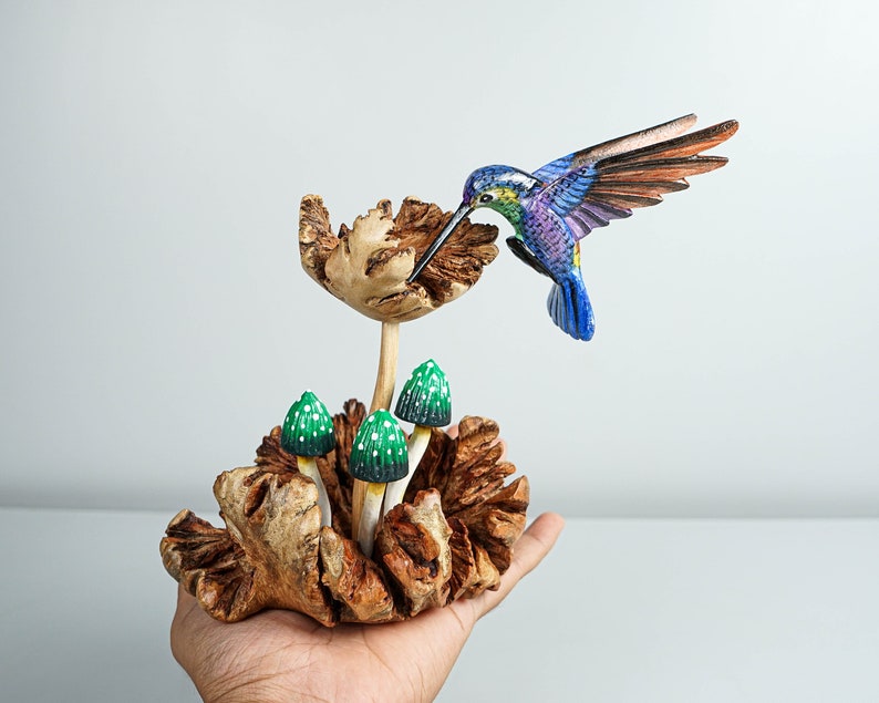 May include: A hand-carved wooden sculpture featuring a vibrant hummingbird with blue, purple, and green plumage hovering over a textured wooden flower, accompanied by three small green mushrooms with white spots emerging from a gnarled wooden base.