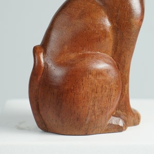 May include: A smooth, polished brown wooden sculpture of a cat in a seated position, showing the side profile of the body, the curved back, and the tail. The wood grain is visible throughout the surface of the figure, which has a warm, medium-brown tone.