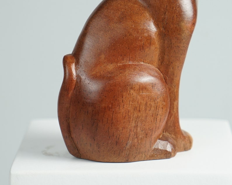 May include: A smooth, polished brown wooden sculpture of a cat in a seated position, showing the side profile of the body, the curved back, and the tail. The wood grain is visible throughout the surface of the figure, which has a warm, medium-brown tone.