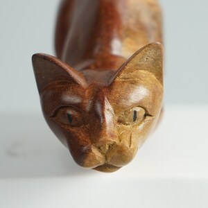 May include: A hand-carved wooden cat figurine featuring a two-tone finish with dark brown wood on the left side and lighter tan wood on the right side. The sculpture displays carved facial features, including pointed ears, almond-shaped eyes, and a nose.