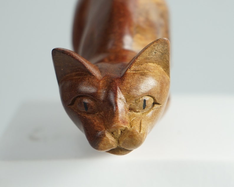 May include: A hand-carved wooden cat figurine featuring a two-tone finish with dark brown wood on the left side and lighter tan wood on the right side. The sculpture displays carved facial features, including pointed ears, almond-shaped eyes, and a nose.