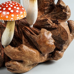 May include: A handcrafted wooden sculpture featuring several mushrooms. One mushroom has a red cap with white spots, while others are white and beige. The mushrooms are set in a piece of textured, brown wood.