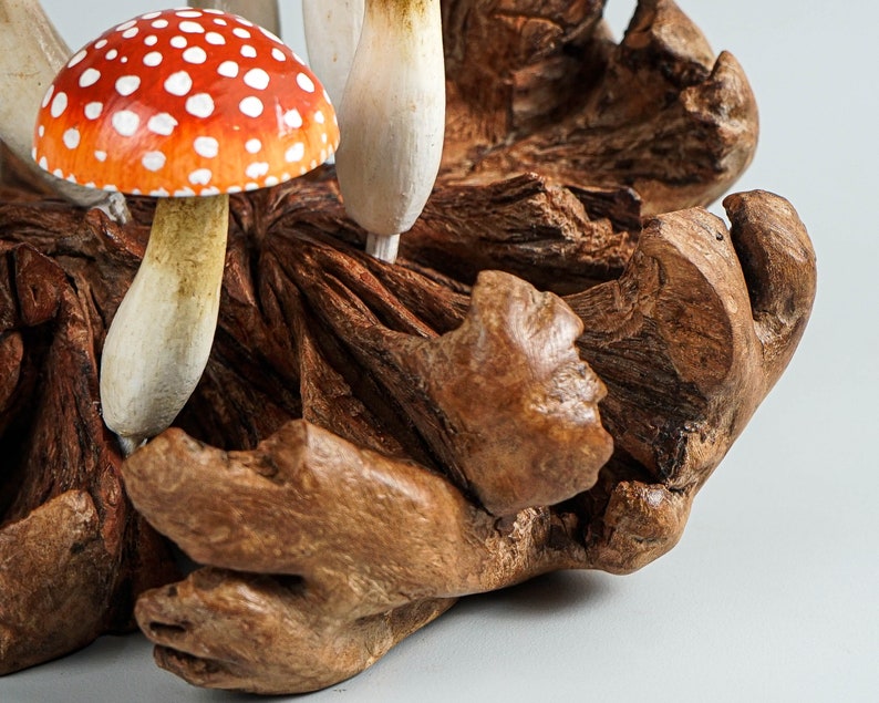 May include: A handcrafted wooden sculpture featuring several mushrooms. One mushroom has a red cap with white spots, while others are white and beige. The mushrooms are set in a piece of textured, brown wood.