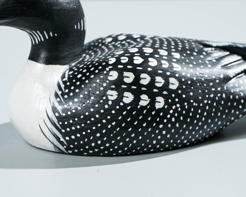 May include: A carved wooden loon figurine featuring a solid black head and neck with white throat markings. The body is painted black with a detailed pattern of white dots and small white feather-like shapes across the back and wings.