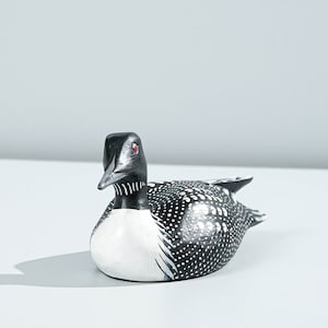 May include: A hand-painted wooden loon figurine featuring a black head, a white chest, and a black back covered in white polka dots. The bird has a sharp, pointed beak and a small, bright red eye, capturing the distinct markings of a common loon.