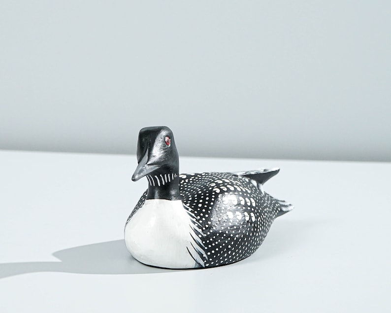 May include: A hand-painted wooden loon figurine featuring a black head, a white chest, and a black back covered in white polka dots. The bird has a sharp, pointed beak and a small, bright red eye, capturing the distinct markings of a common loon.
