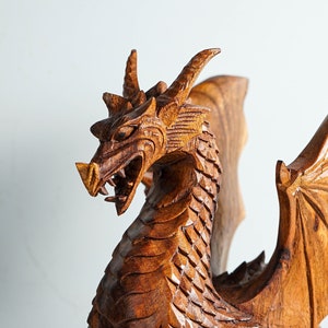 May include: A wooden dragon sculpture with intricate details, featuring a fierce expression and outstretched wings. The dragon is carved from a rich brown wood and has a textured surface.