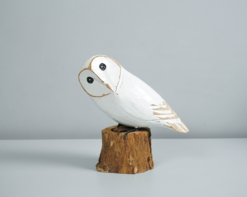 K&ouml;nnte beinhalten: Eine geschnitzte Eulenfigur aus Holz, wei&szlig; bemalt mit schwarzen Augen und braunen Konturen. Die Eule sitzt auf einem kleinen, nat&uuml;rlichen Holzstumpf. Die Fl&uuml;gel der Eule haben ein dezentes, strukturiertes Design.