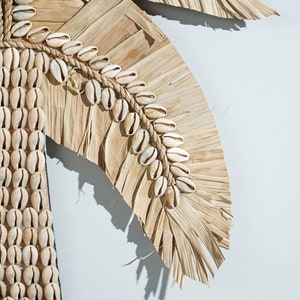 May include: A wall hanging made of dried palm leaves and white seashells. The leaves are arranged in a fan shape and the shells are strung together with twine.
