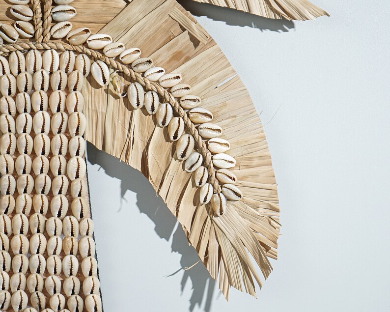 May include: A wall hanging made of dried palm leaves and white seashells. The leaves are arranged in a fan shape and the shells are strung together with twine.