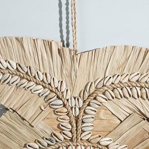 May include: A wall hanging made of natural materials, featuring a woven design of dried palm leaves and cowrie shells. The shells are arranged in a circular pattern and are held together with a rope.