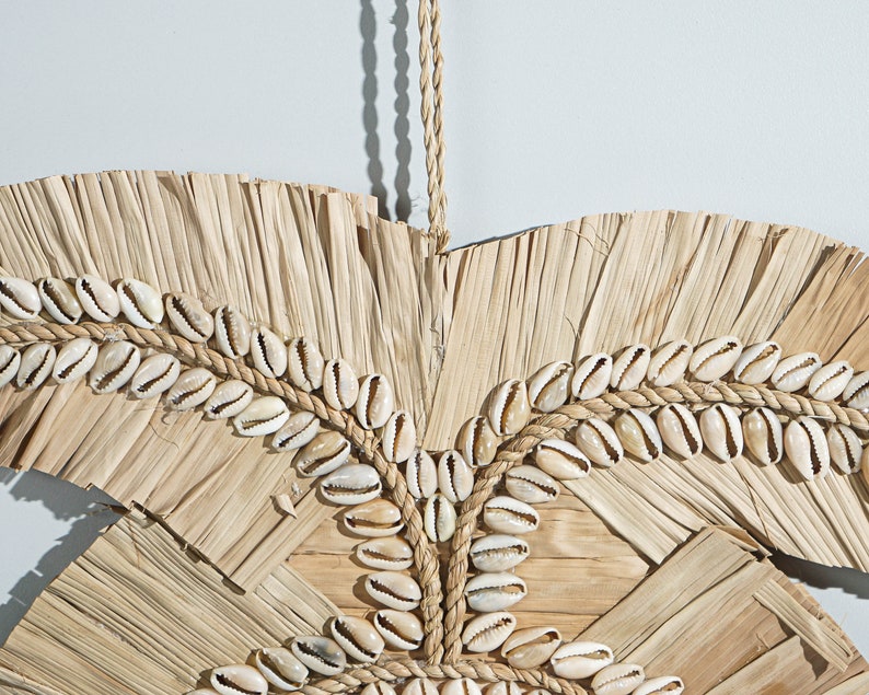 May include: A wall hanging made of natural materials, featuring a woven design of dried palm leaves and cowrie shells. The shells are arranged in a circular pattern and are held together with a rope.