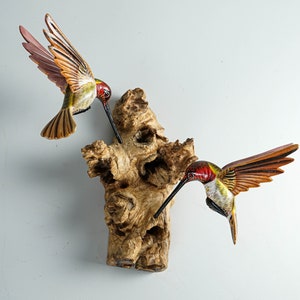 Puede incluir: Dos esculturas de colibr&iacute;es de madera con alas detalladas y largos picos est&aacute;n posadas sobre una base de madera texturizada de color marr&oacute;n claro. Las aves tienen marcas rojas, verdes y amarillas. El fondo es gris claro liso.