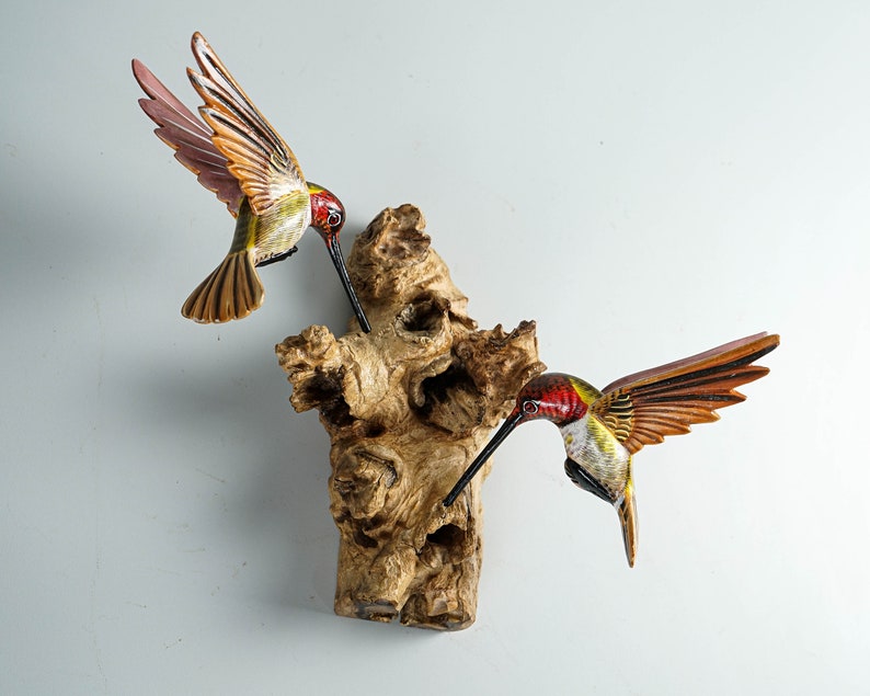 Puede incluir: Dos esculturas de colibr&iacute;es de madera con alas detalladas y largos picos est&aacute;n posadas sobre una base de madera texturizada de color marr&oacute;n claro. Las aves tienen marcas rojas, verdes y amarillas. El fondo es gris claro liso.