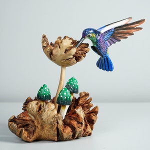 May include: A hand-carved wooden sculpture featuring a vibrant blue and green hummingbird hovering over a textured wooden flower. Three small green mushrooms with white spots are nestled at the base of the sculpture, which is crafted from natural wood.
