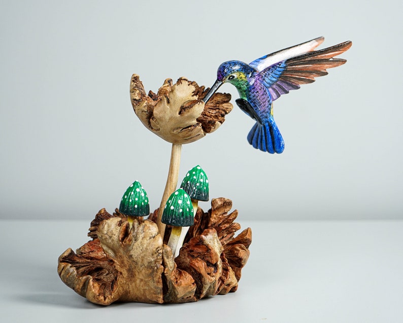 May include: A hand-carved wooden sculpture featuring a vibrant blue and green hummingbird hovering over a textured wooden flower. Three small green mushrooms with white spots are nestled at the base of the sculpture, which is crafted from natural wood.