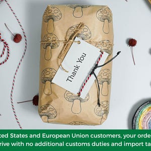 May include: A rectangular package wrapped in light brown paper featuring a repeating black line drawing of mushrooms. The package is tied with thin jute twine and includes a white rectangular gift tag with the black printed text Thank You.