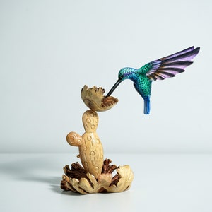 May include: A wooden hummingbird figurine with blue, green, and purple paint, perched on a wooden cactus sculpture. The cactus is made of three pieces, with a base, a middle section, and a top piece that looks like a flower.