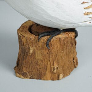 K&ouml;nnte beinhalten: Eine geschnitzte Holzfigur eines Vogels mit wei&szlig; und braun bemalten Details. Die schwarzen F&uuml;&szlig;e des Vogels stehen auf einem kleinen Sockel aus Naturholz. Die Figur ist ein dekorativer Gegenstand.
