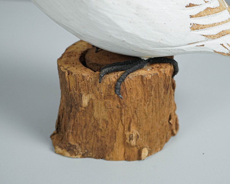 K&ouml;nnte beinhalten: Eine geschnitzte Holzfigur eines Vogels mit wei&szlig; und braun bemalten Details. Die schwarzen F&uuml;&szlig;e des Vogels stehen auf einem kleinen Sockel aus Naturholz. Die Figur ist ein dekorativer Gegenstand.