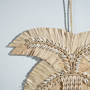 May include: A wall hanging made of woven straw and decorated with white seashells. The hanging is shaped like a palm leaf and has a rope for hanging.