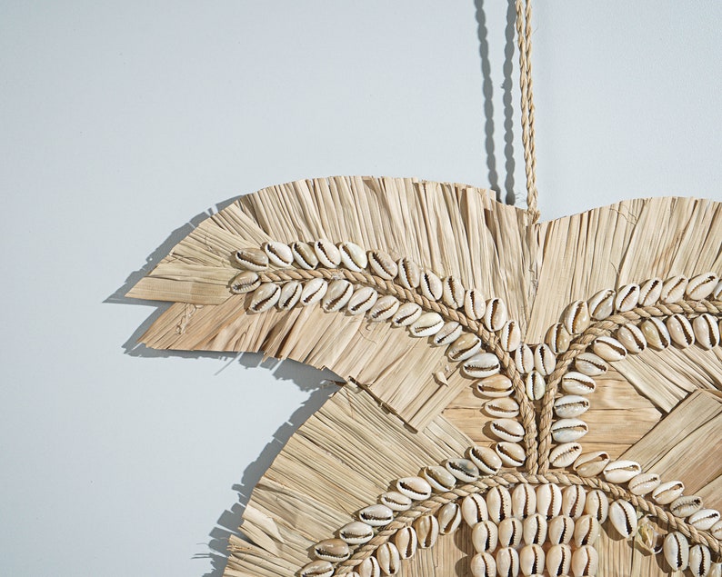 May include: A wall hanging made of woven straw and decorated with white seashells. The hanging is shaped like a palm leaf and has a rope for hanging.