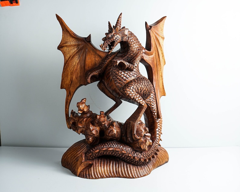 May include: A hand-carved wooden sculpture of a dragon with detailed scales, large wings, and sharp horns. The creature is depicted standing on a textured, rocky base with its tail coiled around the bottom, showcasing intricate wood grain patterns.