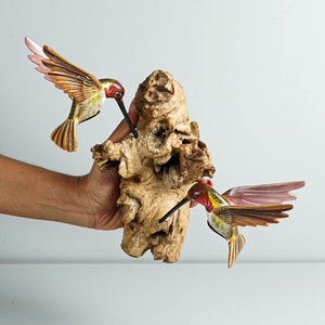 Puede incluir: Dos esculturas de colibr&iacute;es de madera con detalles rojos, amarillos y marrones est&aacute;n posadas sobre un trozo de madera natural. Los colibr&iacute;es tienen las alas extendidas y largos picos, pareciendo alimentarse.