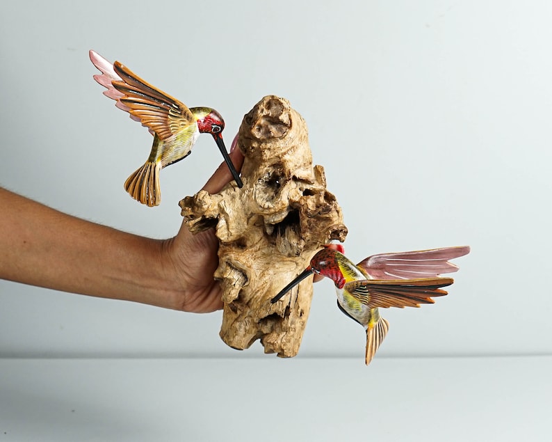 Puede incluir: Dos esculturas de colibr&iacute;es de madera con detalles rojos, amarillos y marrones est&aacute;n posadas sobre un trozo de madera natural. Los colibr&iacute;es tienen las alas extendidas y largos picos, pareciendo alimentarse.