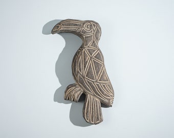 May include: A dark brown wooden toucan wall hanging featuring a geometric pattern of carved lines across its body, head, and beak. The bird is depicted in profile with a large curved beak, a defined eye, and a fan-shaped tail, showcasing a textured finish.