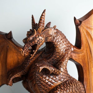 May include: A hand-carved wooden dragon sculpture featuring intricate scale detailing, large spread wings, and a textured head with horns. The figure is crafted from a medium-brown wood with a polished finish, showing the natural grain of the material.