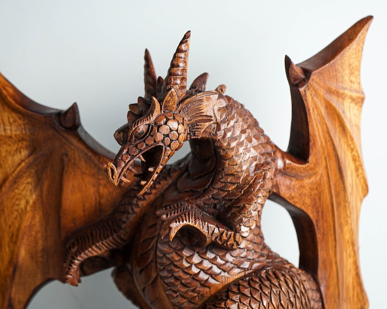 May include: A hand-carved wooden dragon sculpture featuring intricate scale detailing, large spread wings, and a textured head with horns. The figure is crafted from a medium-brown wood with a polished finish, showing the natural grain of the material.