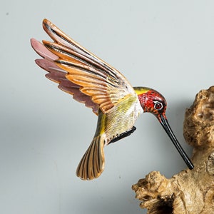 Puede incluir: Una escultura de colibr&iacute; de madera tallada a mano con las alas extendidas, pintada con colores vibrantes. El ave presenta una cabeza roja, detalles amarillos y verdes, y alas naranjas y rosas. Est&aacute; posada cerca de un trozo de madera natural.
