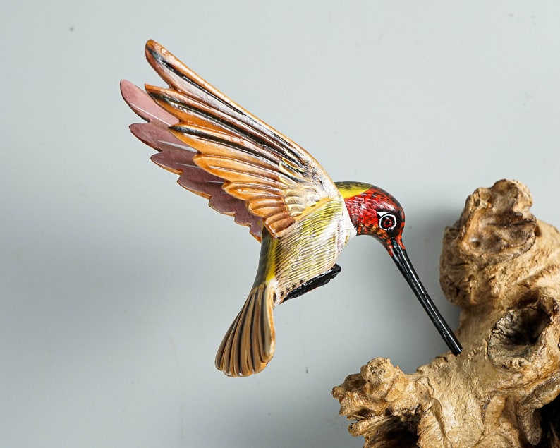 Puede incluir: Una escultura de colibr&iacute; de madera tallada a mano con las alas extendidas, pintada con colores vibrantes. El ave presenta una cabeza roja, detalles amarillos y verdes, y alas naranjas y rosas. Est&aacute; posada cerca de un trozo de madera natural.