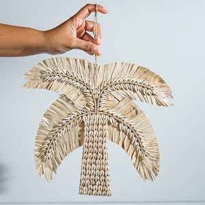 May include: A woven palm tree wall hanging with white seashells. The palm tree is made of natural materials and has a rustic look.