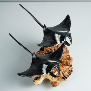 May include: Two black and white manta ray sculptures mounted on a wooden base. The rays are in a flying position with their wings spread out.