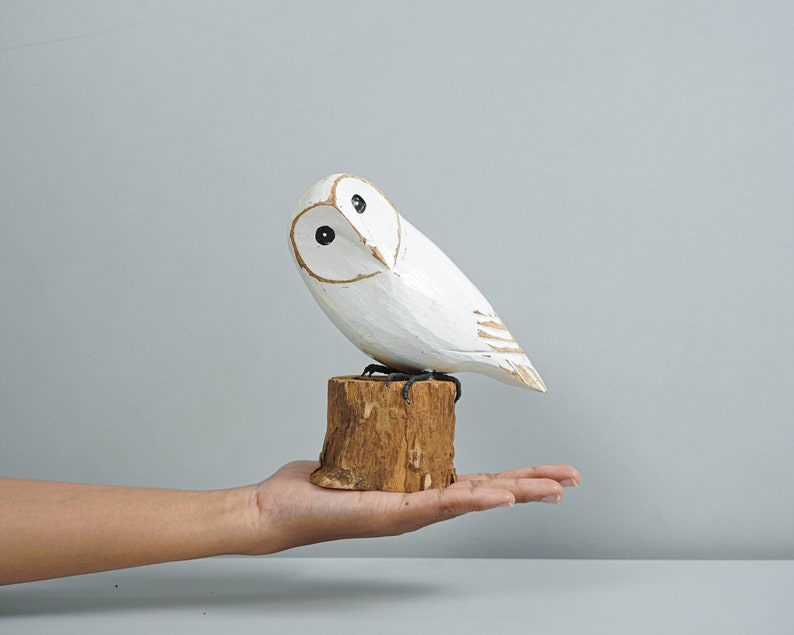 K&ouml;nnte beinhalten: Eine wei&szlig;e Holz-Eulenfigur mit schwarzen Augen und goldenen Akzenten, die auf einem kleinen, nat&uuml;rlichen Holzstumpf sitzt. Die Eule ist handgeschnitzt und hat eine rustikale, strukturierte Oberfl&auml;che. Ein dekorativer Artikel f&uuml;r die Inneneinrichtung.