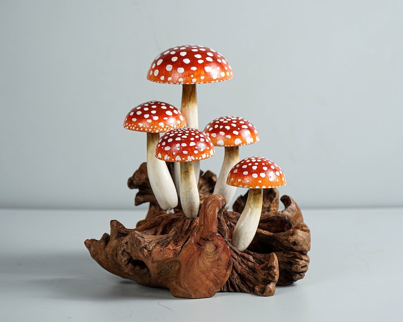 May include: A cluster of five decorative mushrooms with red caps and white spots. The mushrooms have white stems and are attached to a piece of natural wood. The wood has a dark brown color and a textured surface.