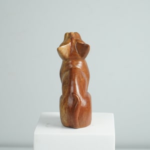 May include: A hand-carved wooden figurine of a seated animal, viewed from the back. The sculpture features a smooth, polished finish with natural brown wood grain patterns, pointed ears, and a curved tail resting against the base of the body.