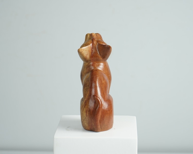 May include: A hand-carved wooden figurine of a seated animal, viewed from the back. The sculpture features a smooth, polished finish with natural brown wood grain patterns, pointed ears, and a curved tail resting against the base of the body.