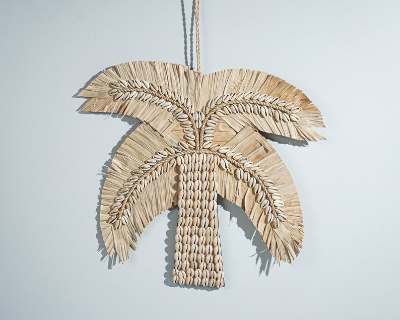 May include: A wall hanging in the shape of a palm tree made from natural materials. The leaves are made from woven straw and the trunk is made from white seashells.